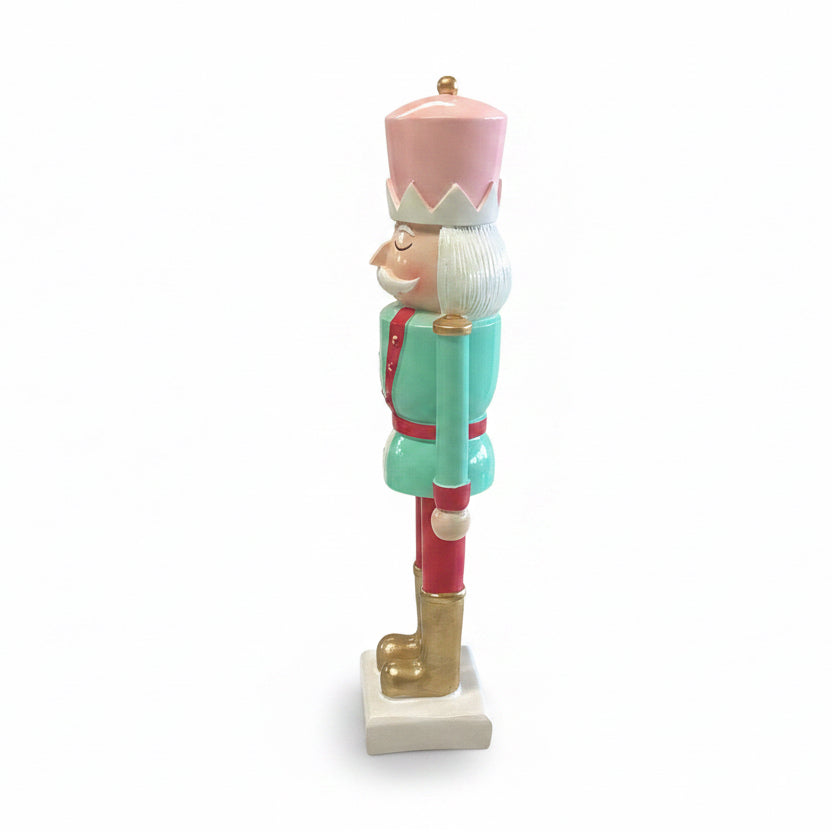 Side view of a Holiday Nutcracker figurine in a pink hat, mint green jacket with red trim, and gold boots.