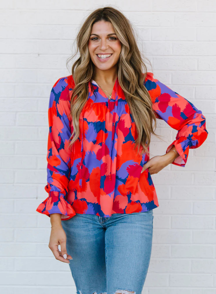 A woman with long, wavy hair smiling and posing in front of a white brick wall, wearing the Elena Midnight Haze Orange top paired with ripped blue jeans.