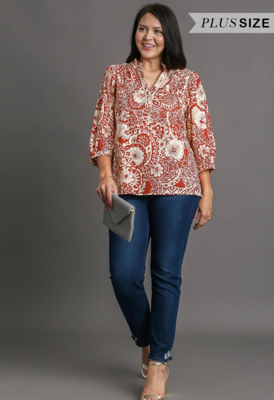 A smiling woman in a Clay Mixed Print Top Plus and jeans, holding a clutch, on a gray background with a "plus size" label in the top right corner.