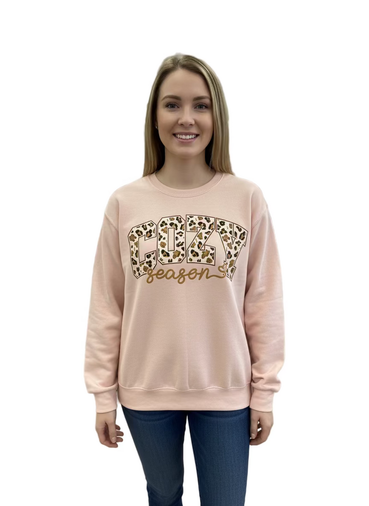 Light pink crewneck sweatshirt with leopard print “COZY” and “season” in cursive. Available in various sizes.