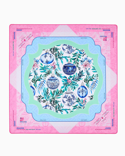 Merriment Mat in Tube: festive table mat with pink border, blue and green inner frames, central floral pattern with blue and white ornaments, and designated playable card areas. Available sizes: standard and large.
