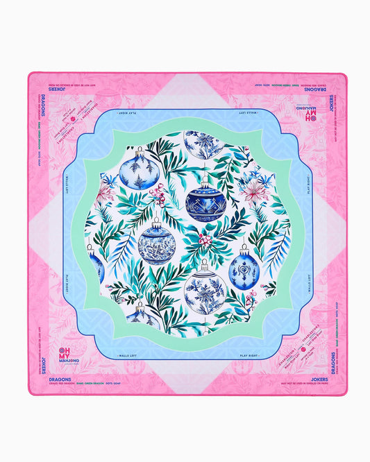 Merriment Mat in Tube: festive table mat with pink border, blue and green inner frames, central floral pattern with blue and white ornaments, and designated playable card areas. Available sizes: standard and large.