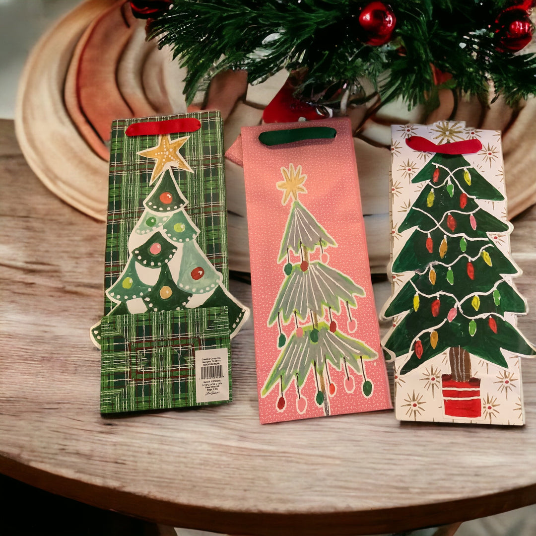 Three tall, rectangular Christmas gift bags with die-cut tree designs in green plaid, pink, and white with ribbon handles.
