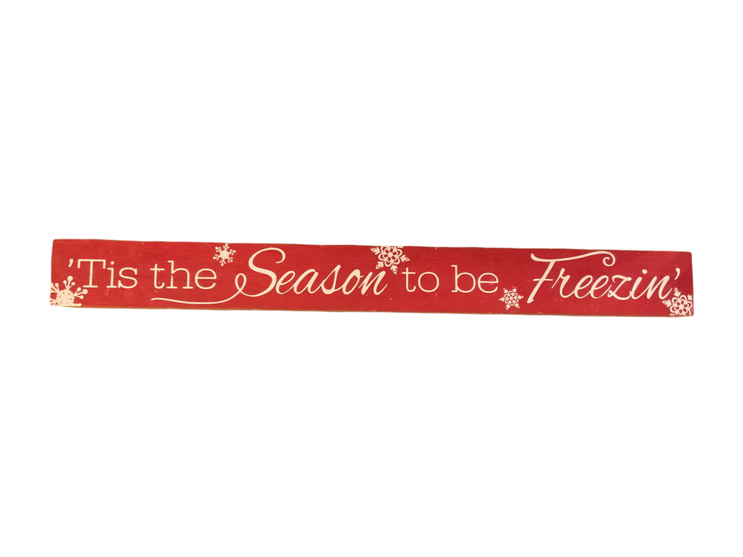 Long, rectangular red wood Christmas sign with white snowflake accents and the text, “‘Tis the Season to be Freezin’.”.