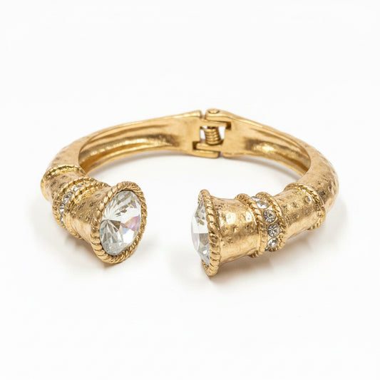 The Jojo Gold Clear Crystal Hinge Bangle features a gold textured design with two large, faceted clear crystals at each end, connected by a gold hinge. Available in multiple sizes.
