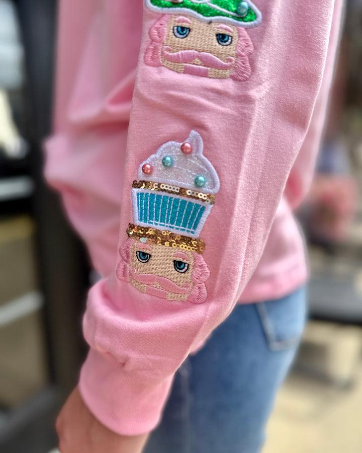 Close-up of a pink Holly Jolly Sweatshirt sleeve with festive sequin cupcake and mustached character patches. Available in sizes S-XL.