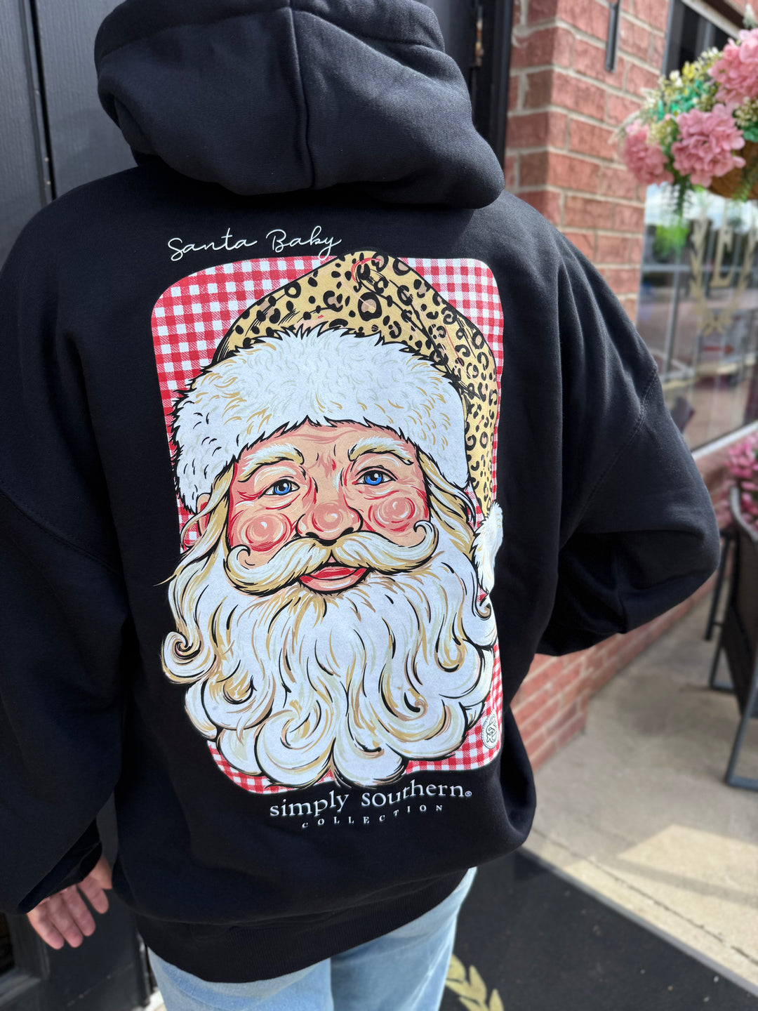 Black Merry Vibes Santa Hoodie with back graphic of Santa Claus in a leopard-print hat on a red and white checkered background, “Santa Baby” text, and brand details. Available in all sizes.