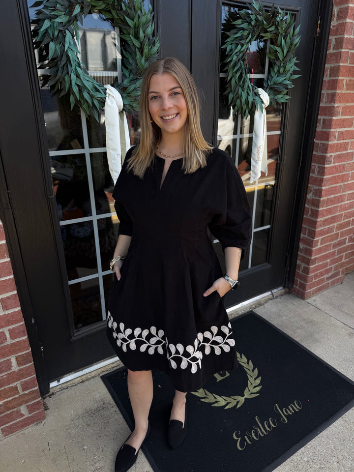 Black corduroy midi dress with white vine embroidery at the hem and a split neckline. Available in various sizes.