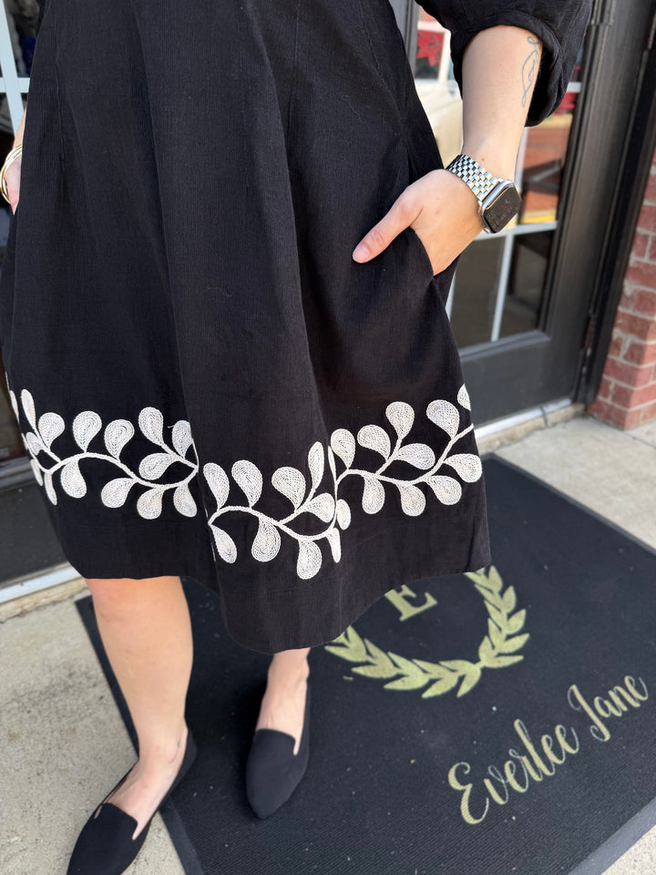 Black corduroy Vine Embroidered dress with white vine embroidery along the hem and side pockets.