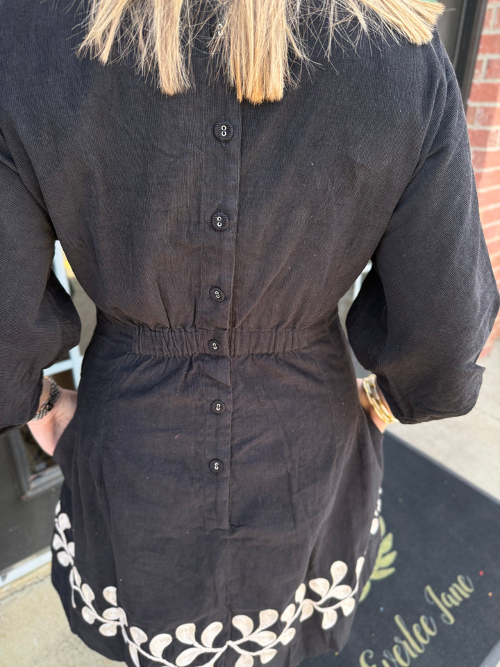 Black corduroy Vine Embroidered Dress with button-up back, elasticized waist, and white hem embroidery. Available in all sizes.