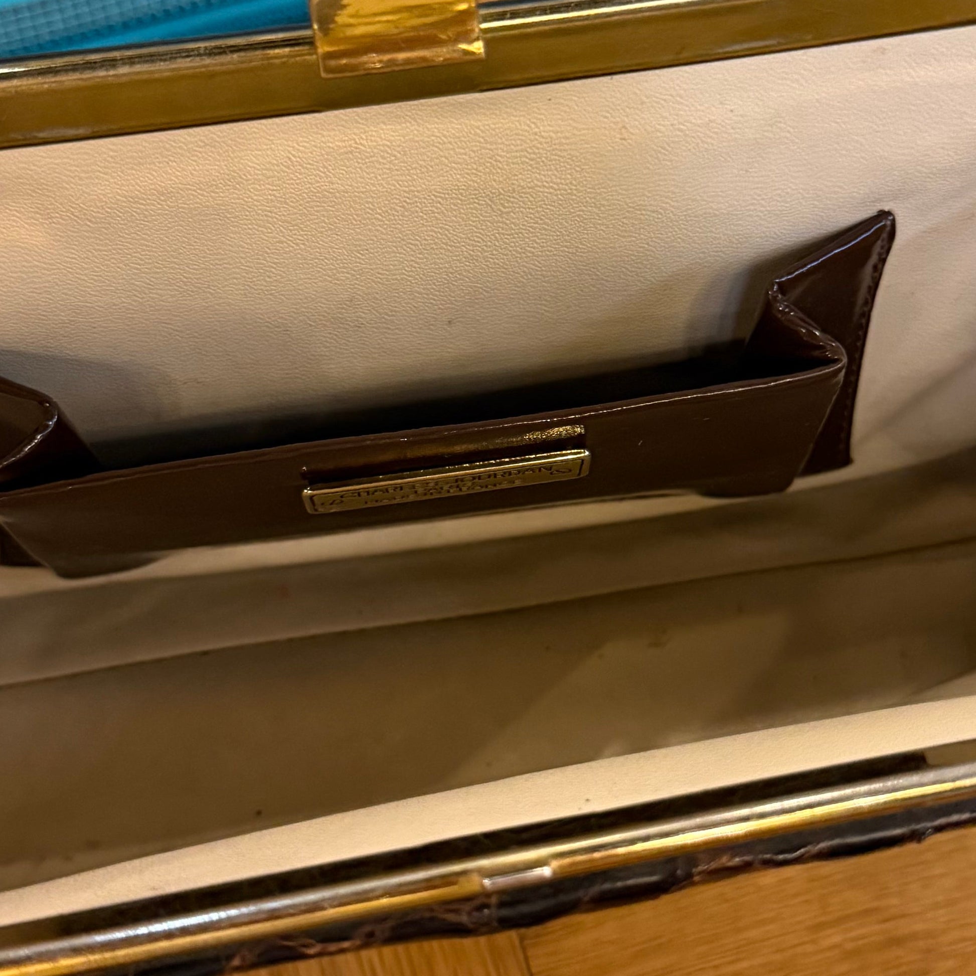 Interior of vintage Charles Jourdan Paris alligator clutch with light lining, brown leather pocket, metallic nameplate, and gold frame. Available size not specified.