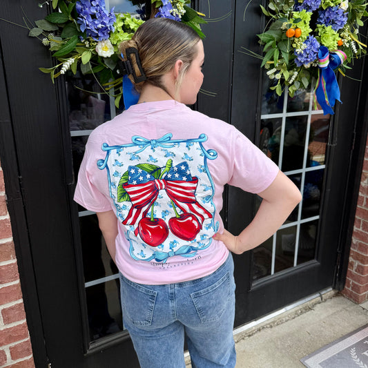 USA Cherry Bow SS T-Shirt in peony pink, patriotic cherries and flags on back. Available in sizes XS–XL.