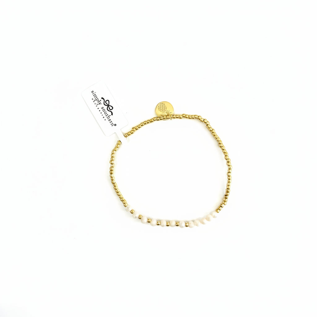 Gold beaded bracelets with white accent beads, round gold charm, and "tiny things" tag. Sizes available: S, M, L.