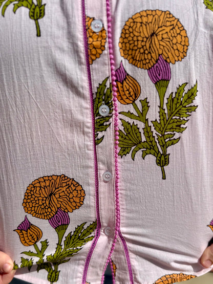White cotton button-up blouse with yellow, green, purple florals and pink-stitched button line. Available in XS–XL.