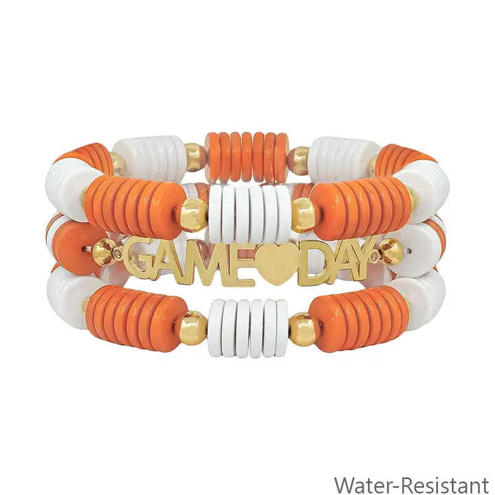 Three beaded stretch bracelets in orange, white, and gold, each with a central gold plate engraved "GAME♥DAY," featuring round and disc-shaped beads. Water-resistant. Available in multiple sizes.