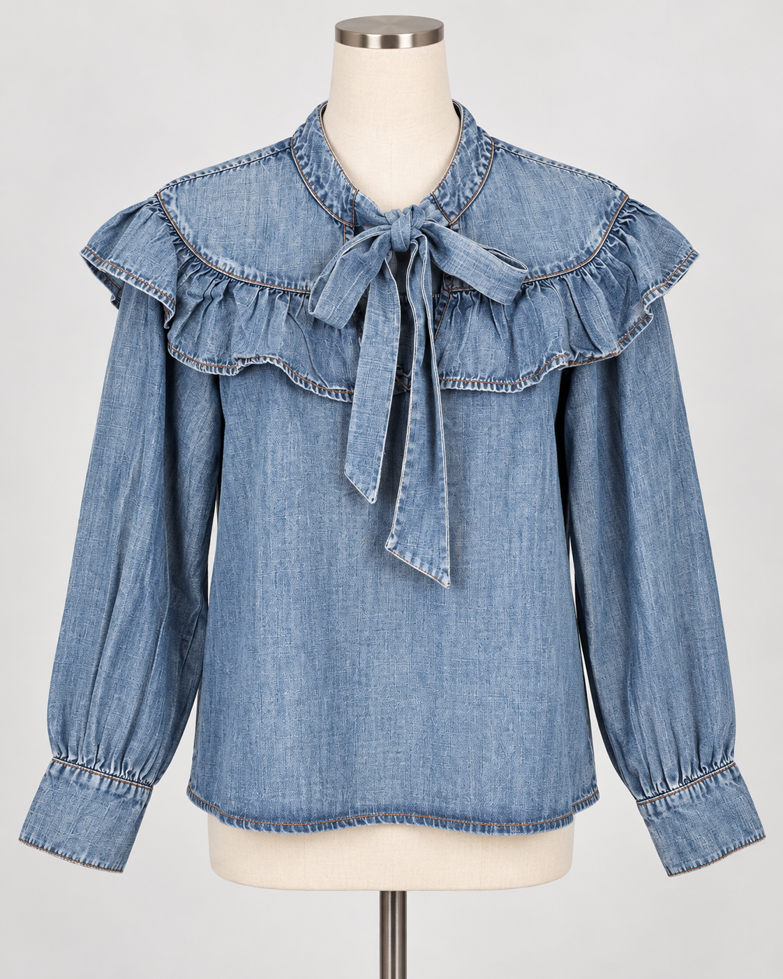 The Denim Bow Top is a light blue Tencel denim blouse with long sleeves, a ruffled yoke, tie-neck bow, and gathered cuffs. Available in sizes XS–XL.