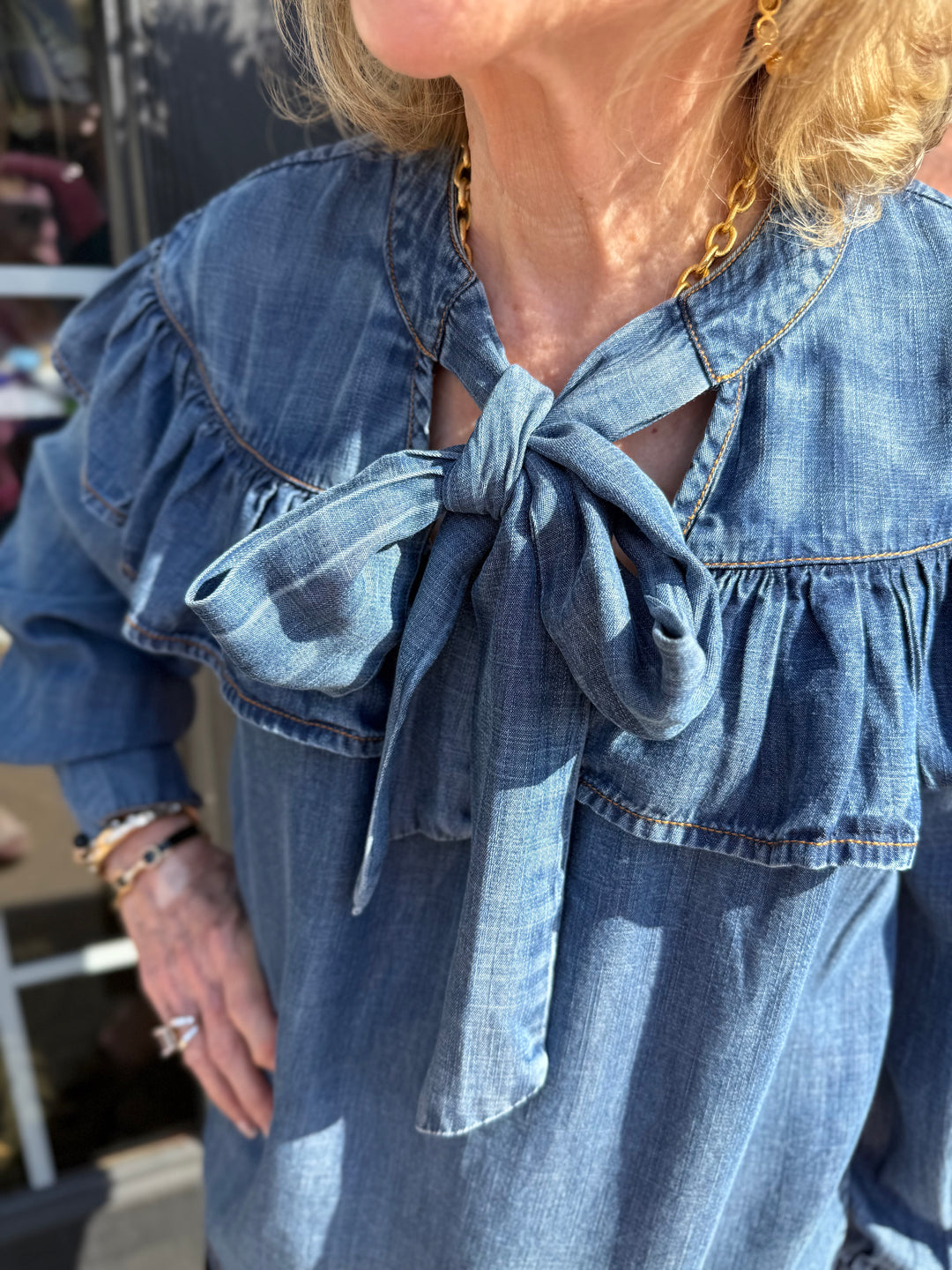 The Denim Bow Top is a Tencel blouse featuring a large collar bow and ruffled detailing. Available in sizes XS-XL.