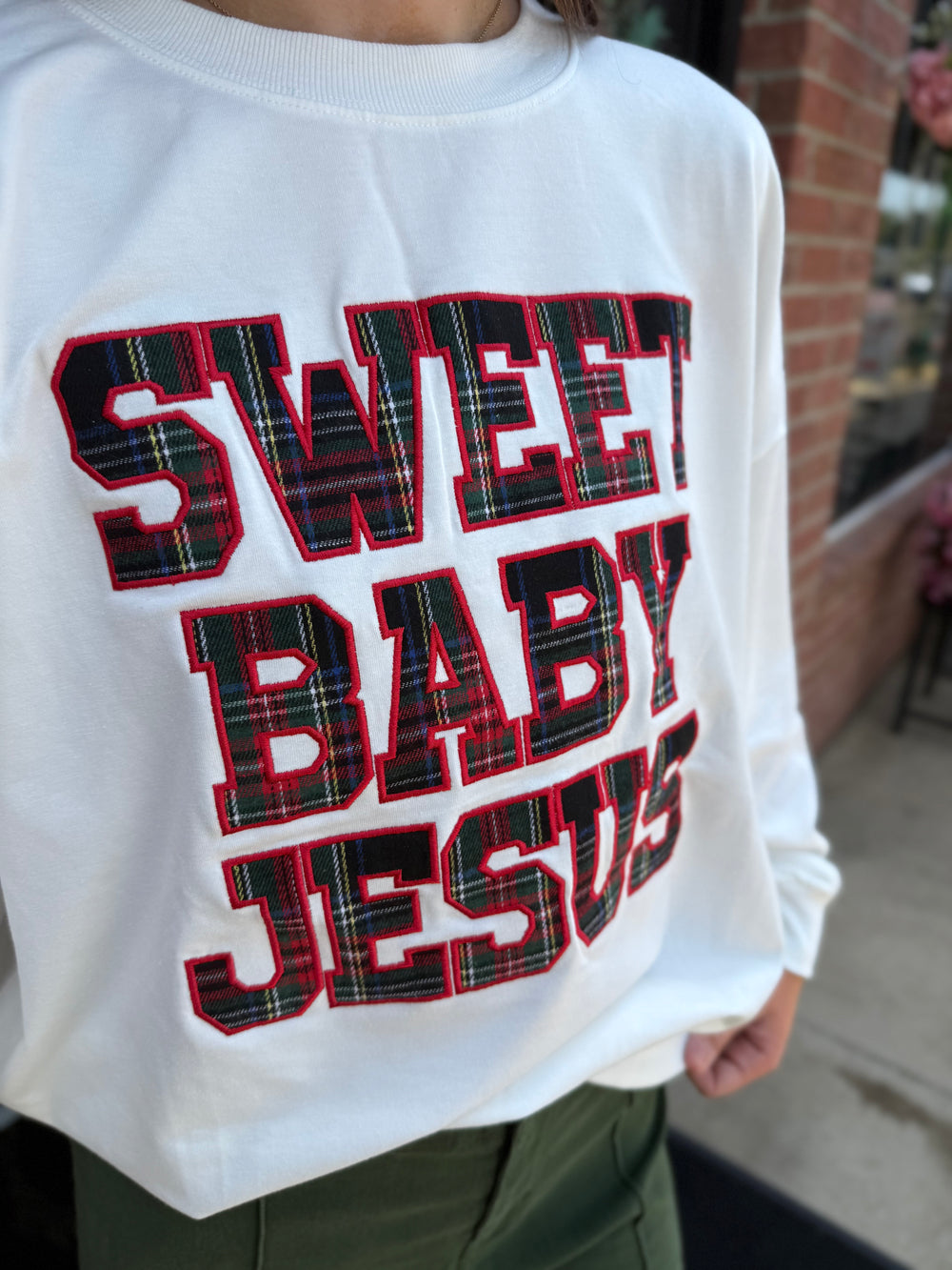 White long-sleeve Sweet Baby Jesus crew neck sweatshirt with plaid “SWEET BABY JESUS” lettering and red outline.