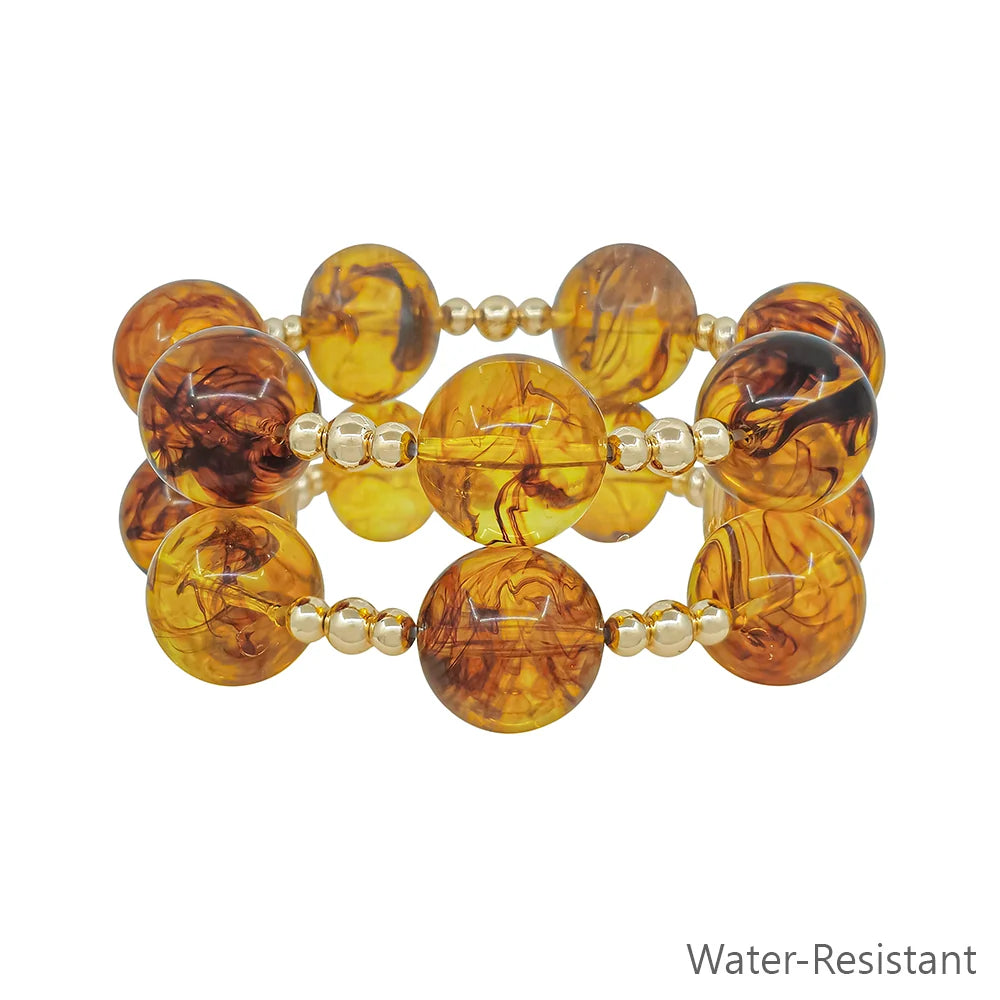 Jet Tortoise Stretch Bracelet Set features two rows of round amber beads with swirling patterns, separated by small gold beads. Stretchy and water-resistant. Sizes available: S, M, L. "Water-Resistant" text in gray at bottom right.