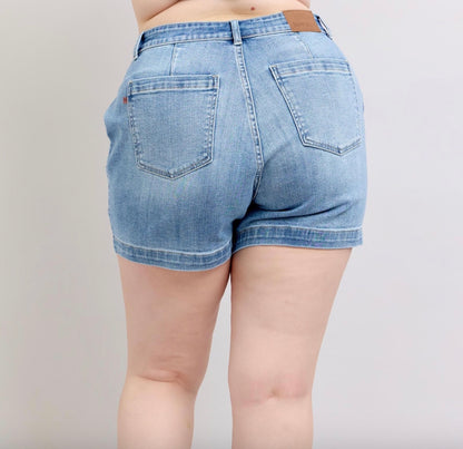 Judy Blue Mid Rise Trouser Shorts in light blue feature visible stitching, back pockets, and a leather patch on the waistband. Available sizes: XS to XL.