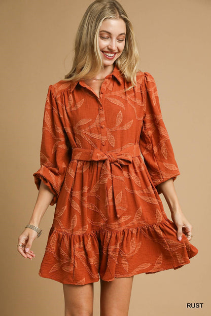 Rust-colored Piper Jacquard Dress with leaf pattern, tie waist, ruffled hem, and buttons. Available in sizes XS-XL.