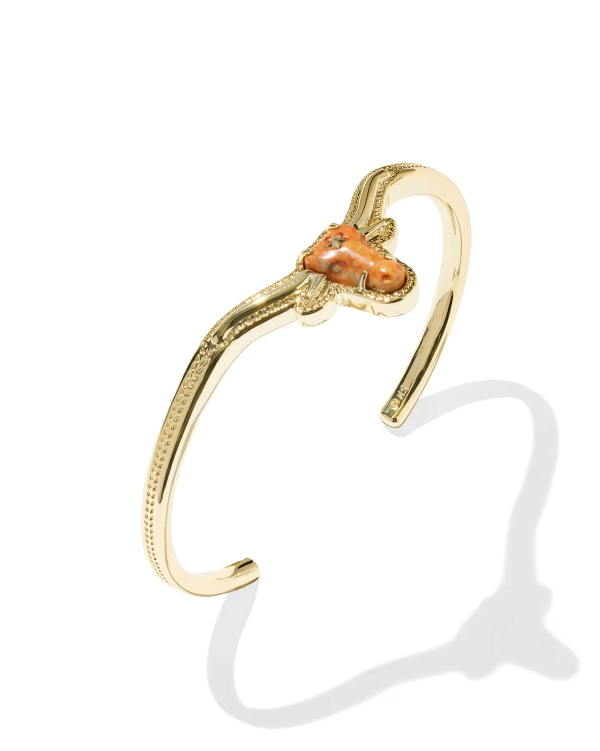 The Longhorn Gold Cuff Bracelet by Kendra Scott features an orange magnesite heart-shaped stone at the center with textured gold detailing on the band. Available in one adjustable size.