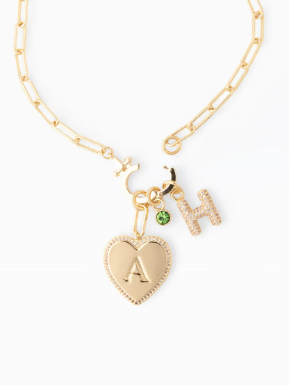 18k gold plated, hypoallergenic Thea Charm Bar necklace with customizable charms: heart-shaped pendant with “A,” crystal-studded “H,” green gem accent, and crescent moon. Available in 16″, 18″, and 20″ chain lengths.