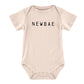 Beige baby onesie with short sleeves and round neck, featuring "NEWBAE" in bold black letters across the chest. Snaps at the bottom for easy diaper changes. Sizes available: Newborn, 0-3 months, 3-6 months.