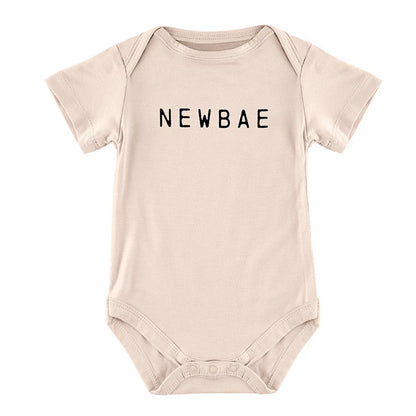 Beige baby onesie with short sleeves and round neck, featuring "NEWBAE" in bold black letters across the chest. Snaps at the bottom for easy diaper changes. Sizes available: Newborn, 0-3 months, 3-6 months.