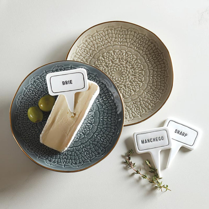 Two decorative plates featuring brie cheese, grapes, and ceramic cheese markers. Includes markers labeled "Manchego" and "Sharp" with thyme. Sizes available: Standard.