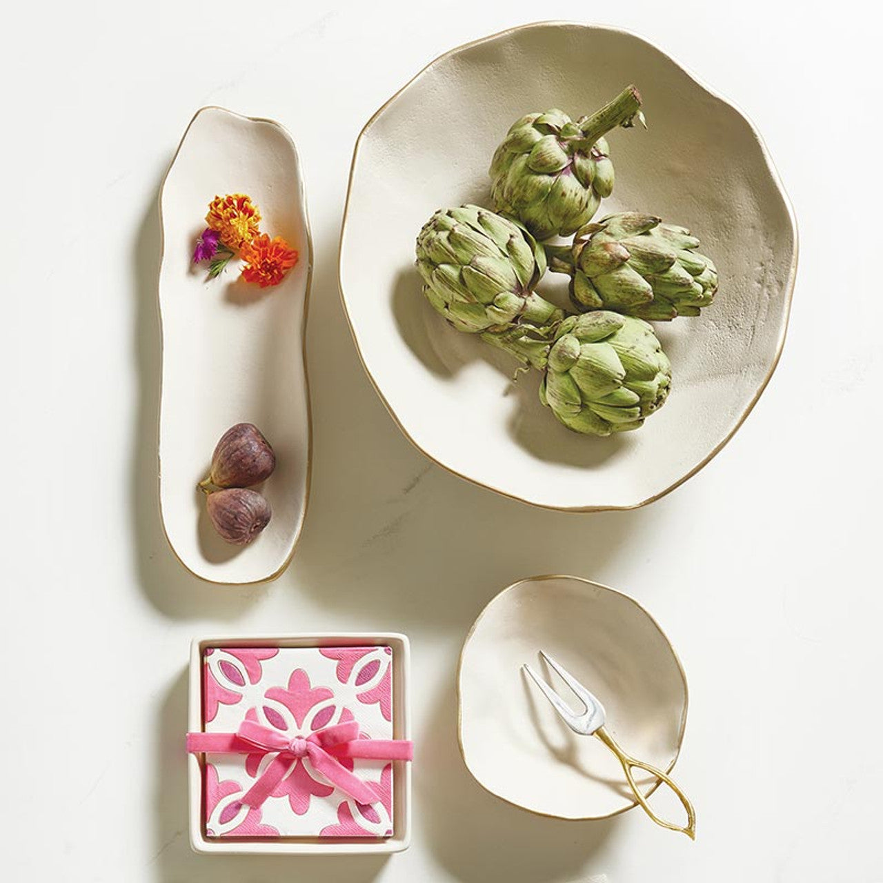 The Ceramic Napkin Tray + Napkins Set includes ceramic plates with artichoke, fig, and floral designs on a white background. A small dish for a decorative knife is included, along with paper cocktail napkins. Available sizes are not specified.