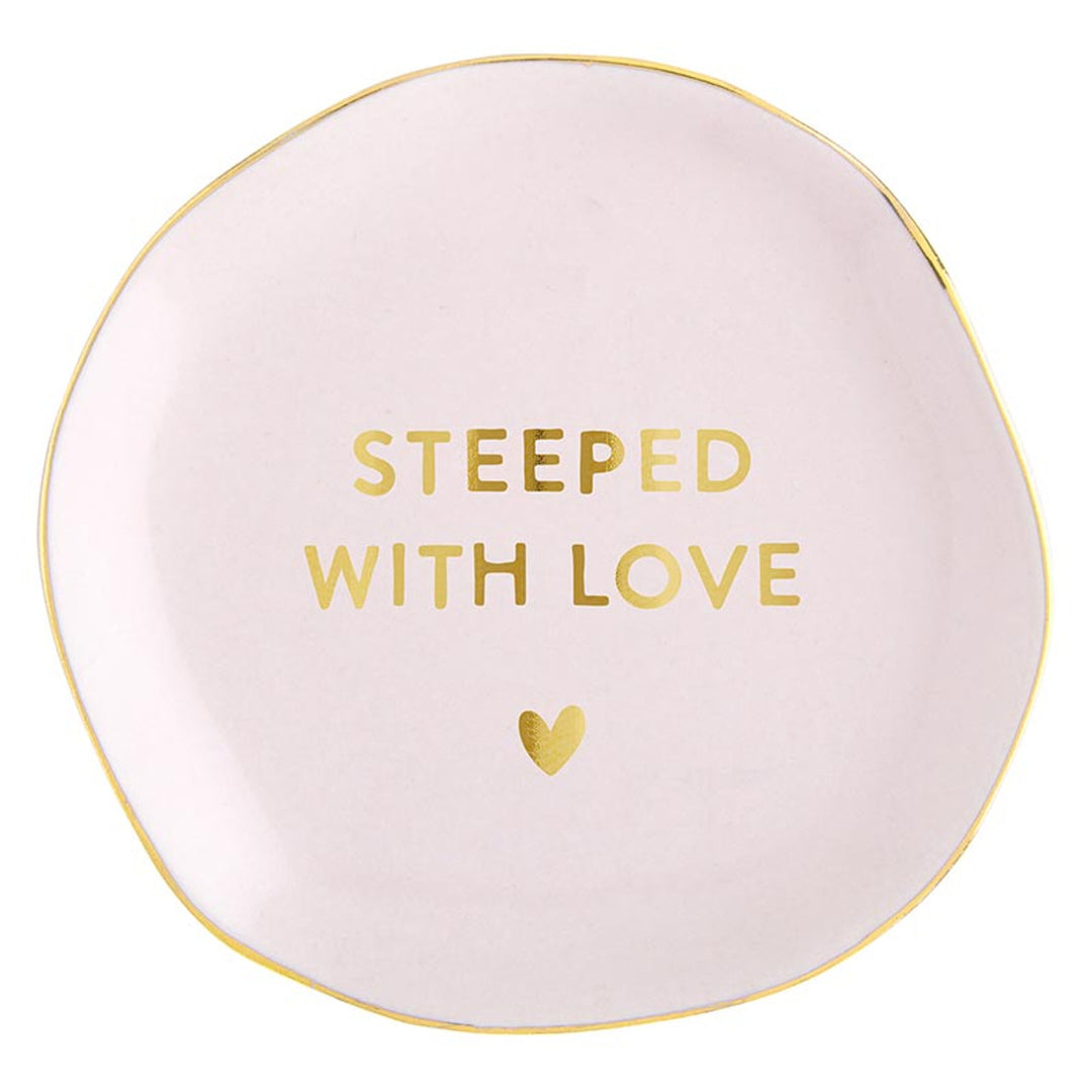 Round, pale pink ceramic tea bag holder with uneven edges and gold trim. Features "STEEPED WITH LOVE" inscription and a small gold heart. Available size: One size.