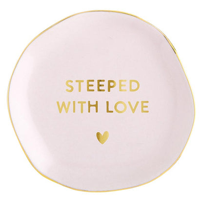 Round, pale pink ceramic tea bag holder with uneven edges and gold trim. Features "STEEPED WITH LOVE" inscription and a small gold heart. Available size: One size.