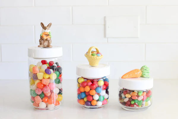 Three Pinstripes Canister jars filled with colorful candies, each with a decorative melamine lid: one topped with a chocolate bunny, one with a basket, and one with a carrot. Sizes available: Small (4" H), Medium (5" H), Large (6" H).