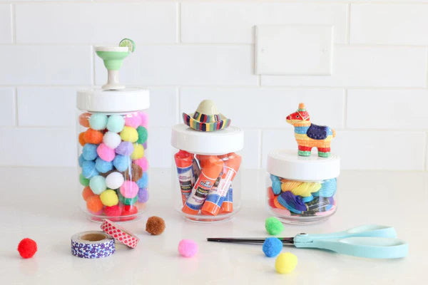 Set of three canisters with melamine lids topped by festive figurines: cocktail glass, sombrero, and piñata. Ideal for storing craft supplies. Sizes available: Small (4" H x 3" Dia), Medium (5" H x 4" Dia), Large (6" H x 5" Dia).