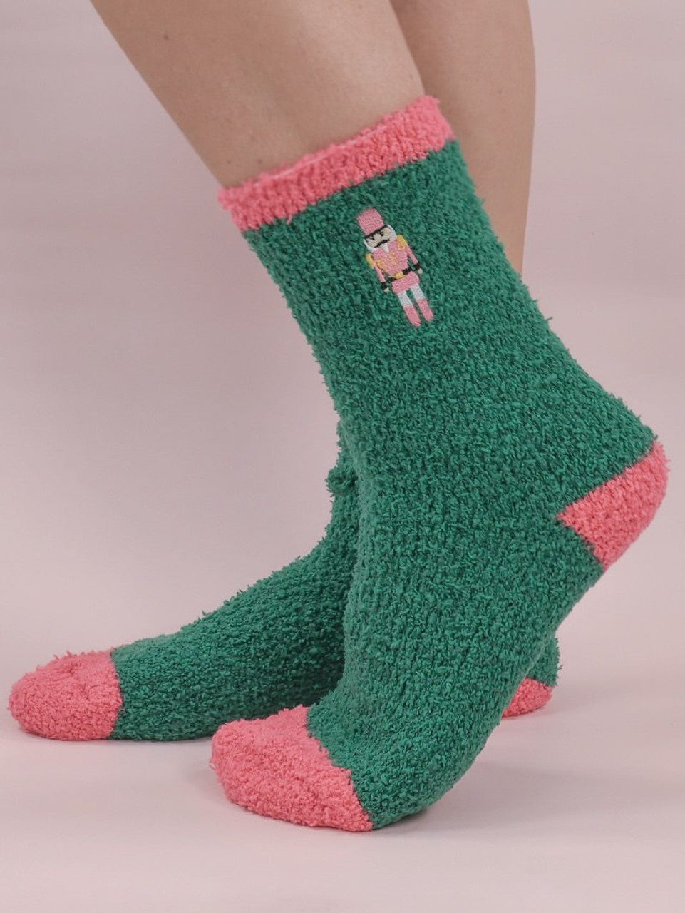 Soft-n-Cozy Fuzzy Holiday Socks are green with pink toes, heels, and cuffs, featuring a small nutcracker design near the top. Available sizes: S/M and M/L.