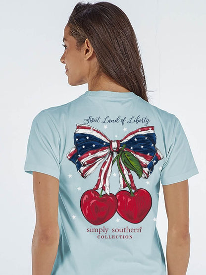 Light blue Simply Southern tee with flag bow, cherries, "Sweet Land of Liberty" on back. Sizes: S-XXL.