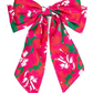 The Holiday Bows feature a large, vibrant bow with a floral pattern in bright pink and green hues, accented with white. It has long, wide tails that showcase the colorful design. Available sizes: Small, Medium, Large.