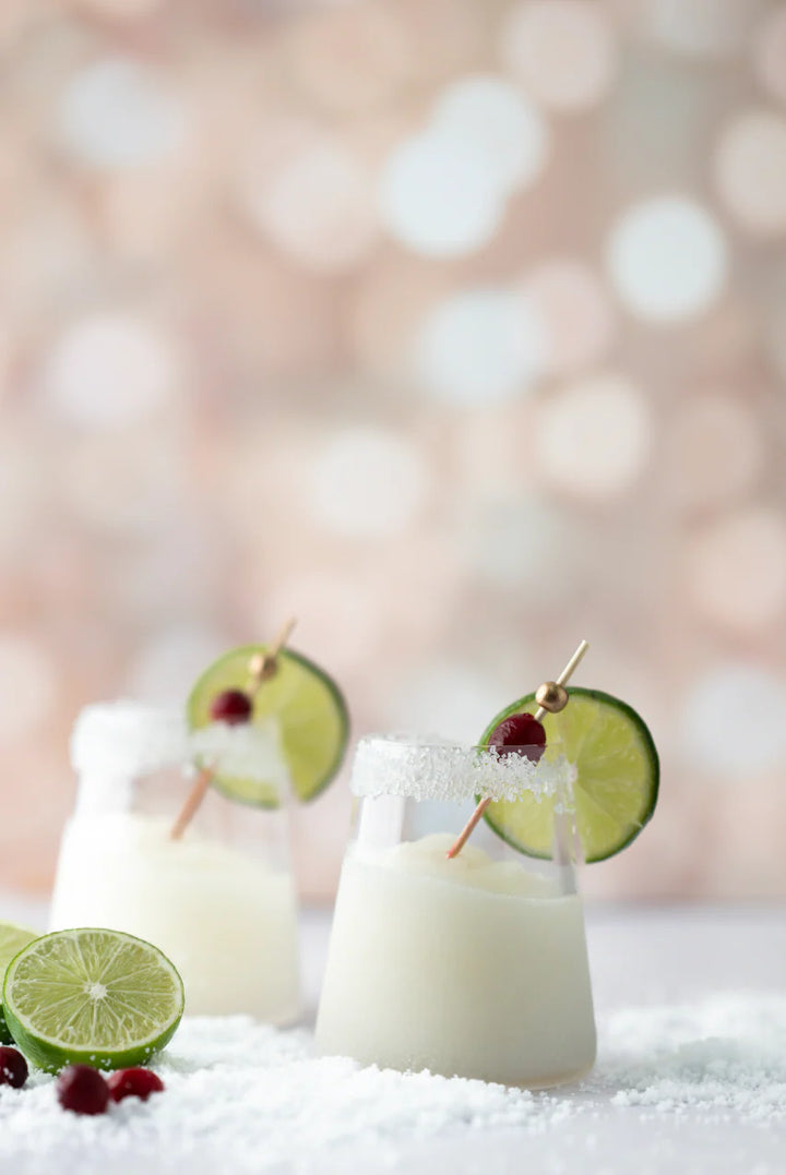 Two glasses of 10 oz. d’marie Drink Mixes White Christmas Margarita, a creamy frozen cocktail rimmed with sugar and garnished with a lime slice and a cranberry on a pick.