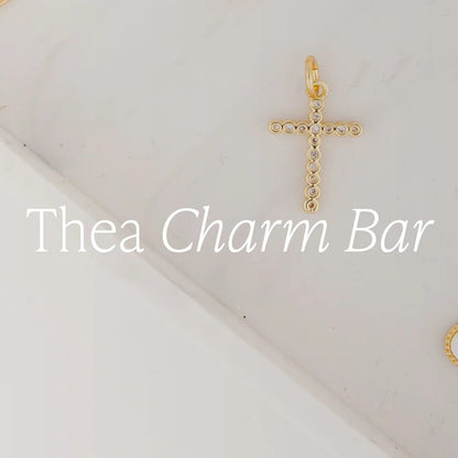 A delicate 18k gold-plated cross pendant with small circular accents, available in multiple sizes. Customizable as part of the Thea Charm Bar collection.