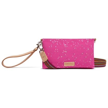 The Uptown Crossbody in Berry is a pink clutch purse with silver speckles, featuring a tan leather wristlet strap, a rainbow-striped shoulder strap, a front tan logo patch, and zipper compartments. Available in small and medium sizes.