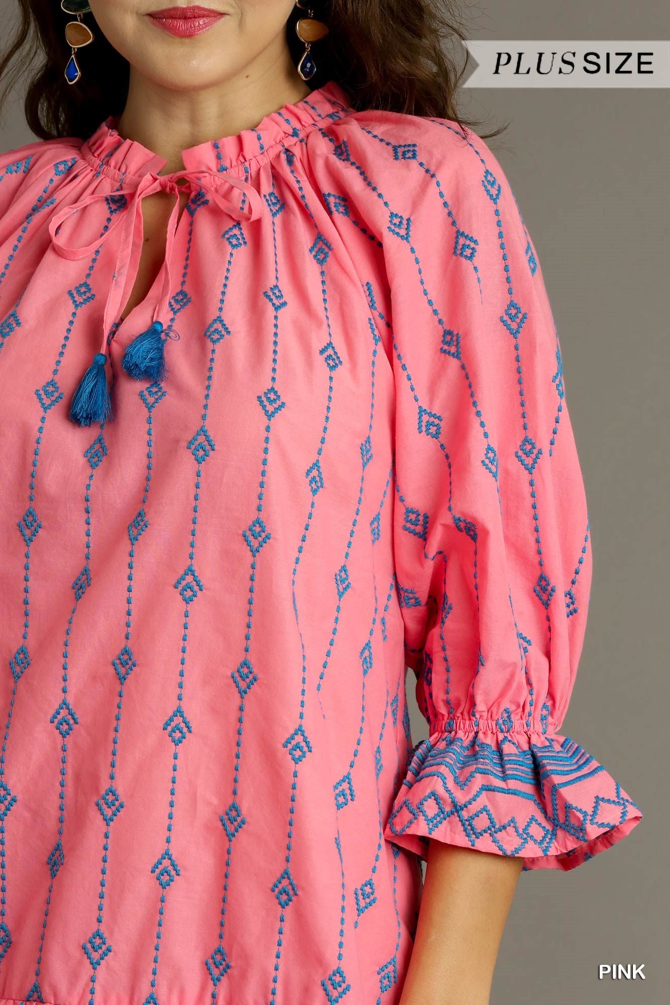 Pink blouse with blue geometric patterns. Features a split ruffle neckline with tassel ties and ruffled sleeves. Short A-Line design with embroidery work. Product available in plus sizes.