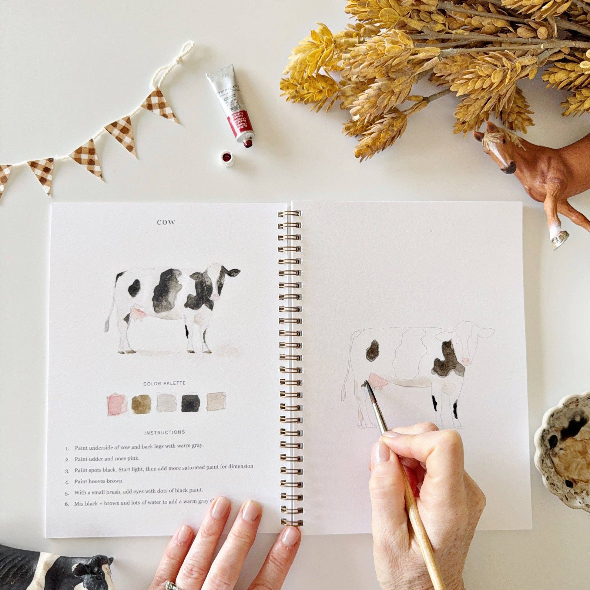 A hand paints a cow in the spiral-bound Farm Watercolor Workbook, which includes painting instructions. Available in 8x10 inches.