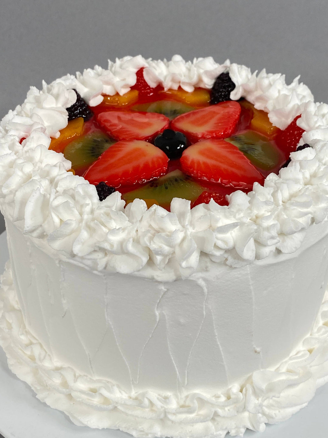 Small round white frosted fake vanilla cake with whipped cream border, topped with faux strawberries, kiwi, blackberries, peaches. Available in small size.