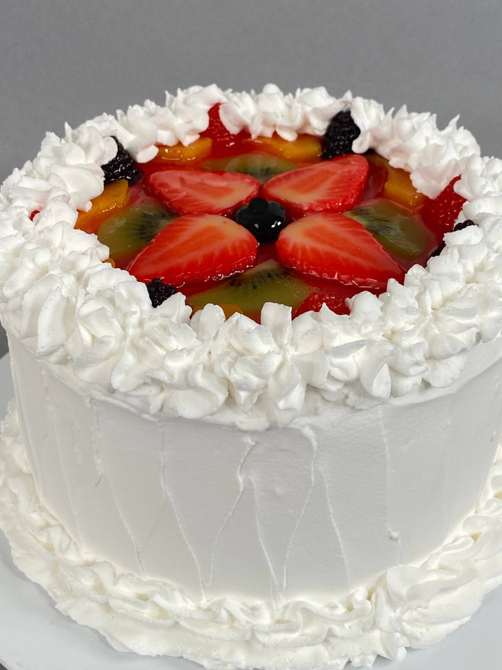 Small round white frosted fake vanilla cake with whipped cream border, topped with faux strawberries, kiwi, blackberries, peaches. Available in small size.