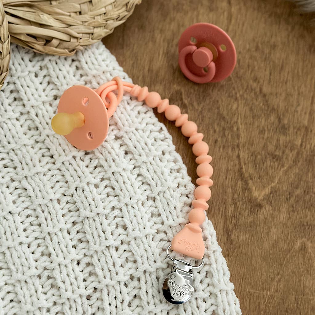 A peach-colored pacifier with a Sweetie Strap™ Silicone One-Piece Pacifier Clips: Mint Braid attached to a metal clip, lying on a textured white blanket next to a basket and a matching cap.