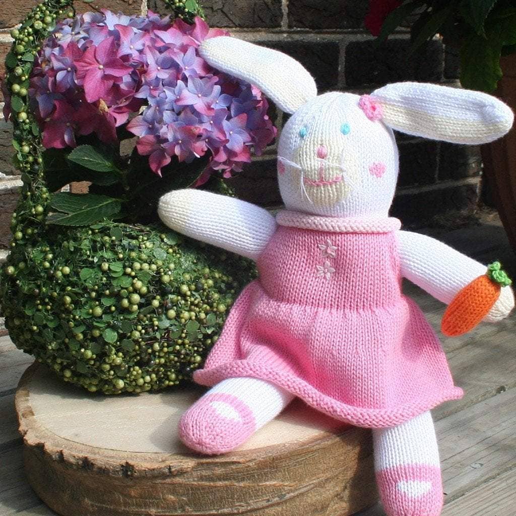 Sentence with product name: Harriett the Bunny Knit Doll: 12" Plush in a pink dress sitting on a stump next to a plant.