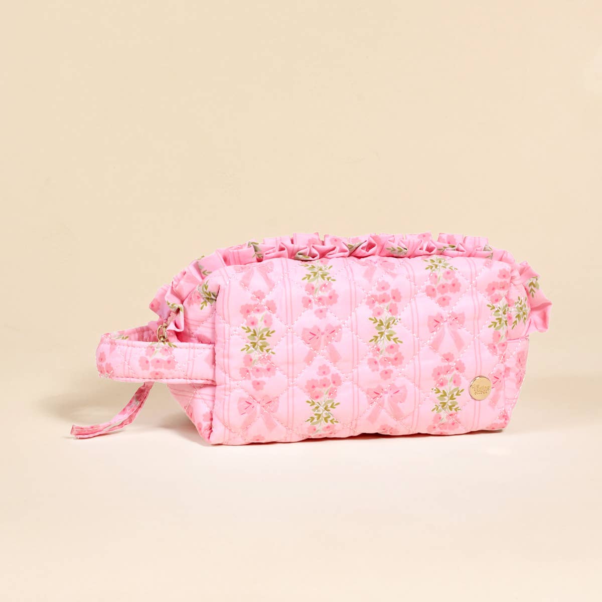 Pink quilted cosmetic pouch with floral and bow prints, ruffled trim, side handle, gold emblem, wipeable interior. Sizes: S/M/L.
