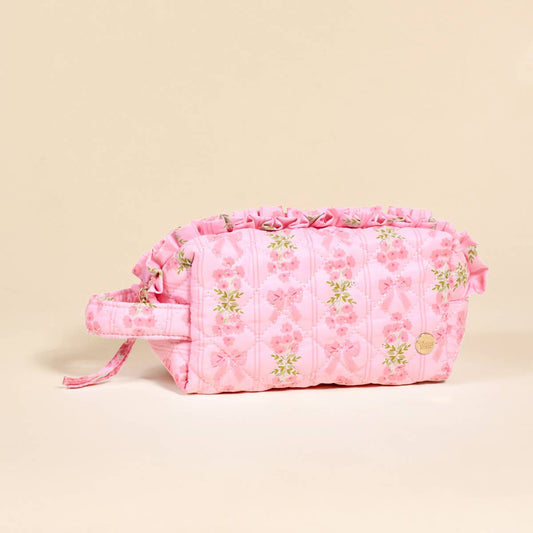 Pink quilted cosmetic pouch with floral and bow prints, ruffled trim, side handle, gold emblem, wipeable interior. Sizes: S/M/L.