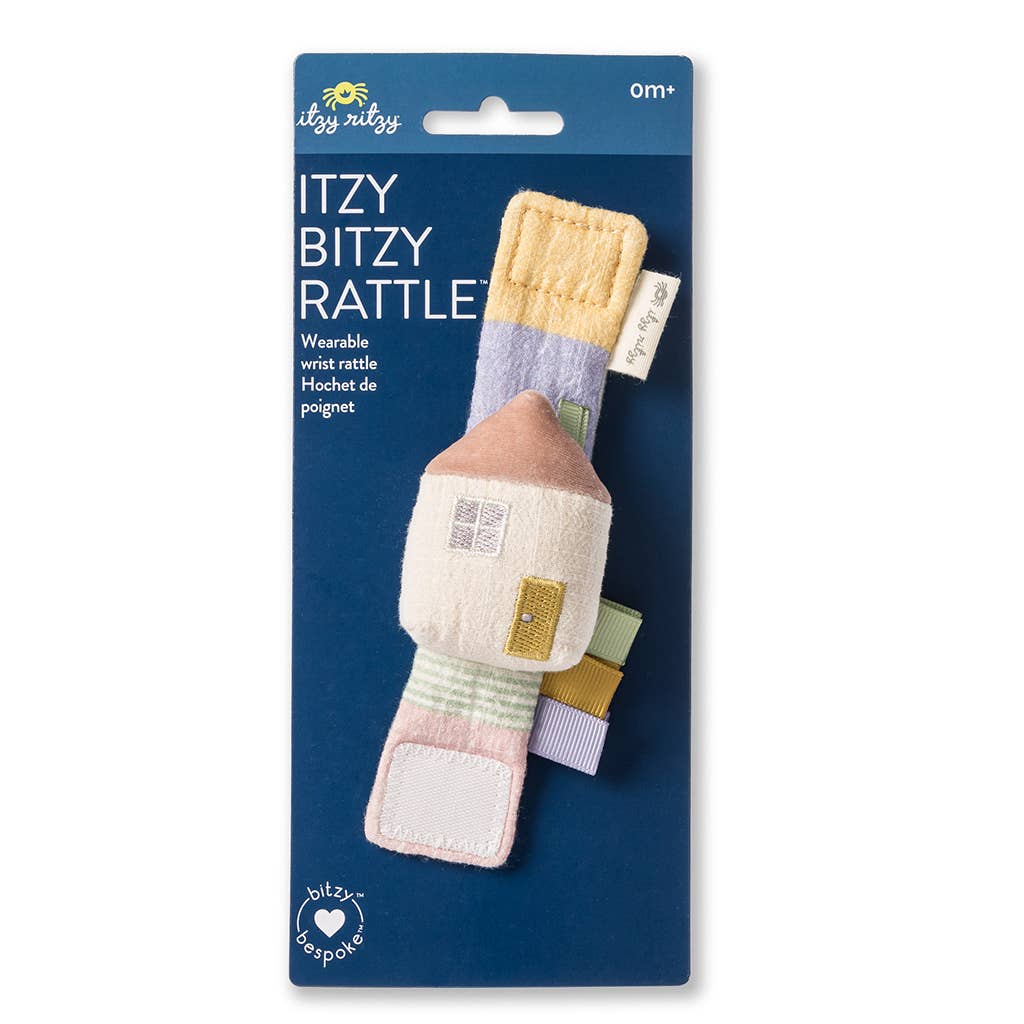 A Itzy Bitzy Wrist Rattle: Cottage for infants, designed to look like a house with natural cotton fabric textures and pastel colors, packaged on a blue card labeled "itzy bitzy rattle.